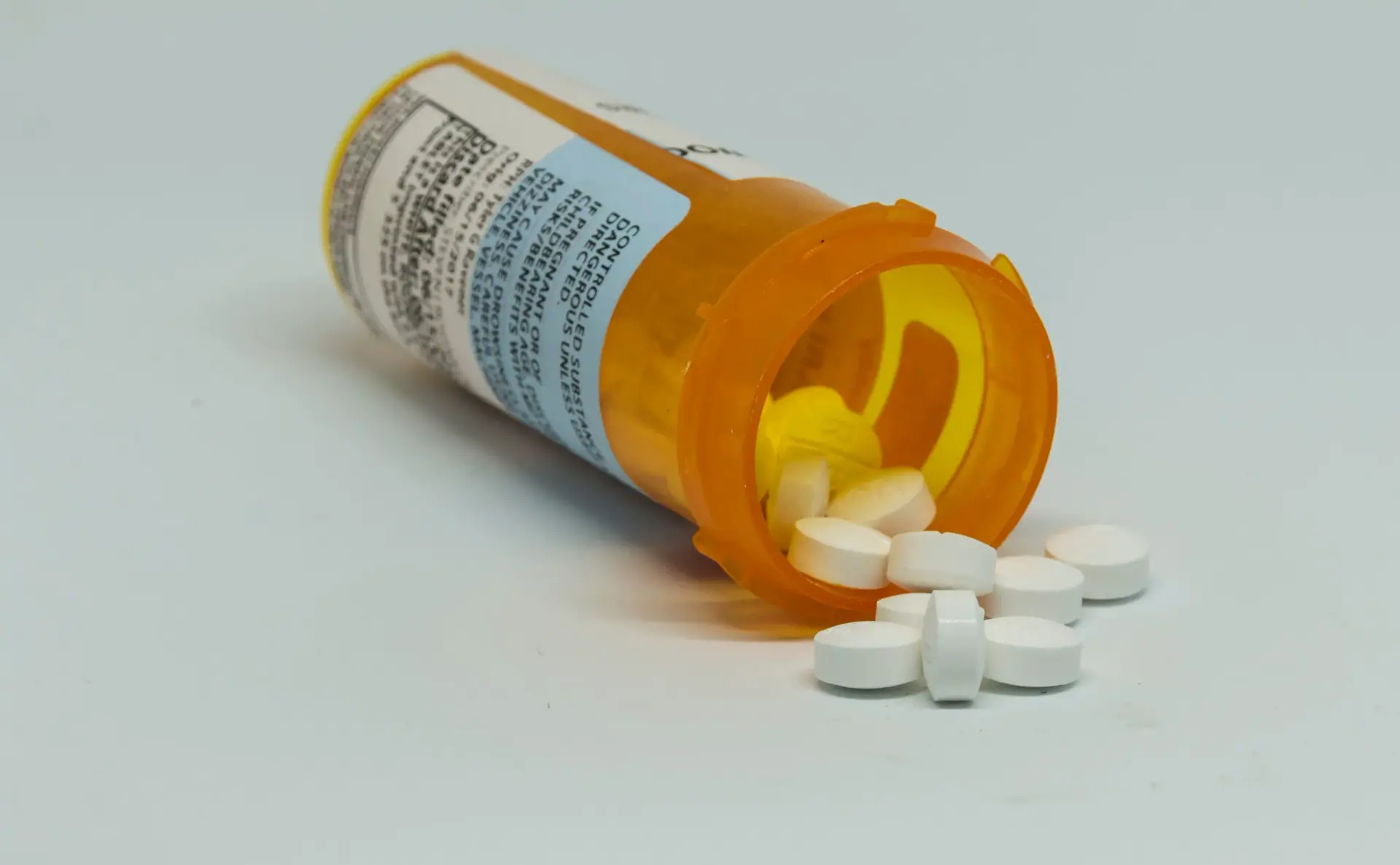 Pill bottle on its side with controlled substance pills falling out of the bottle with a white background.